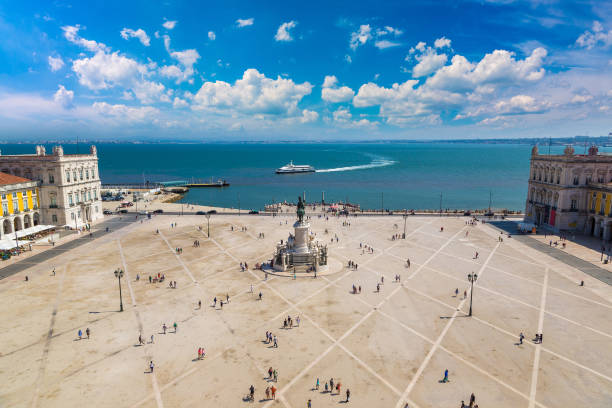 A high-angle view of Praça do Comércio, a large square in Lisbon, Portugal, with a statue of King José I in the center. The square is surrounded by classical buildings with yellow facades, and the Tagus River is visible in the background. A ferry is seen crossing the river, leaving a white wake behind. The sky is blue with scattered white clouds, and people are walking across the square, adding a sense of scale to the scene.