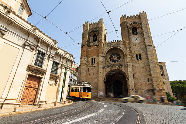 A view of the Lisbon Cathedral (Sé de Lisboa), a historic Romanesque church with two imposing towers and a large rose window in the center. A classic yellow tram is seen curving along the tracks in front of the cathedral, adding a dynamic element to the scene. Overhead tram wires crisscross the sky, while blurred pedestrians and a passing car convey movement. The cobblestone street and surrounding old buildings enhance the historic charm of the setting.