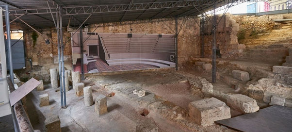 Image of the archaeological excavations of the Roman Theatre of Lisbon, featuring stone ruins and remaining columns. A panel in the background displays a digital reconstruction of the ancient theatre, showing the original layout of the seating and stage. The site is protected by a metal roof structure.