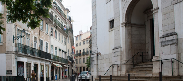 A narrow street in Lisbon, Portugal, lined with colourful buildings. The entrance to the São Cristóvão church is on the right, with a grand archway and stone steps leading up to it.