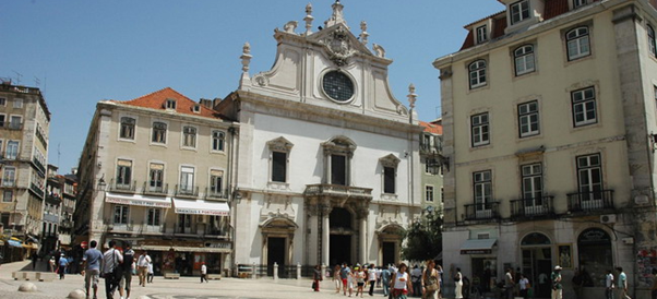 A street-level view of Largo de São Domingos in Lisbon. Traditional Portuguese buildings with painted facades line a narrow street, leading towards the stone steps and arched entrance of the São Cristóvão church on the right. A few people are visible walking along the street, and a white van is parked further down. The church features a grand stone archway and steps leading up to the main entrance