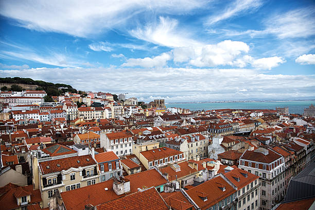 Panoramic view of Lisbon, Portugal, showcasing the city's iconic red-roofed buildings. The image captures the historic Baixa and Alfama districts, with São Jorge Castle and other landmarks visible in the background. The Tagus River stretches towards the horizon, under a bright blue sky with scattered clouds