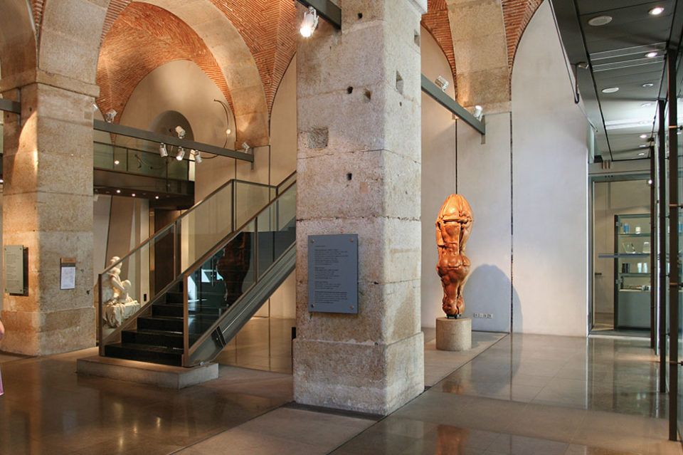 Interior shot of a museum with exposed stone columns, brick arched ceilings, and a contemporary glass and metal staircase. A wooden sculpture is visible in the right foreground.