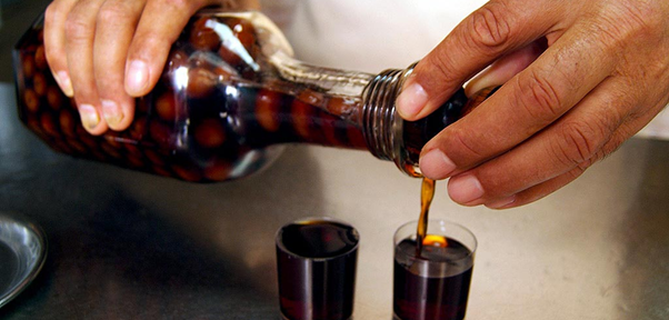 A person pours a dark red liqueur (Gingnha, a very populer Lisbon liqueur) from a glass bottle with a decorative pattern into small plastic cups. The liqueur contains whole cherries, which are visible through the bottle. The setting appears to be a bar or café, with a reflective metal surface underneath.